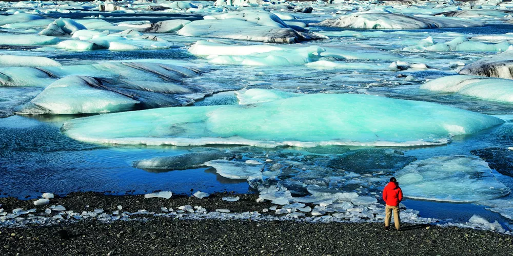 Member of tour in red stood next to the glacial lagoon Lake Jokulsarlon in Iceland