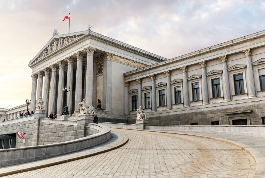 A red and white Austrian flag flies atop Austria's Parliament Building, a large grey building with enormous columns and a v shaped roof, with a curved brick driveway running down. 