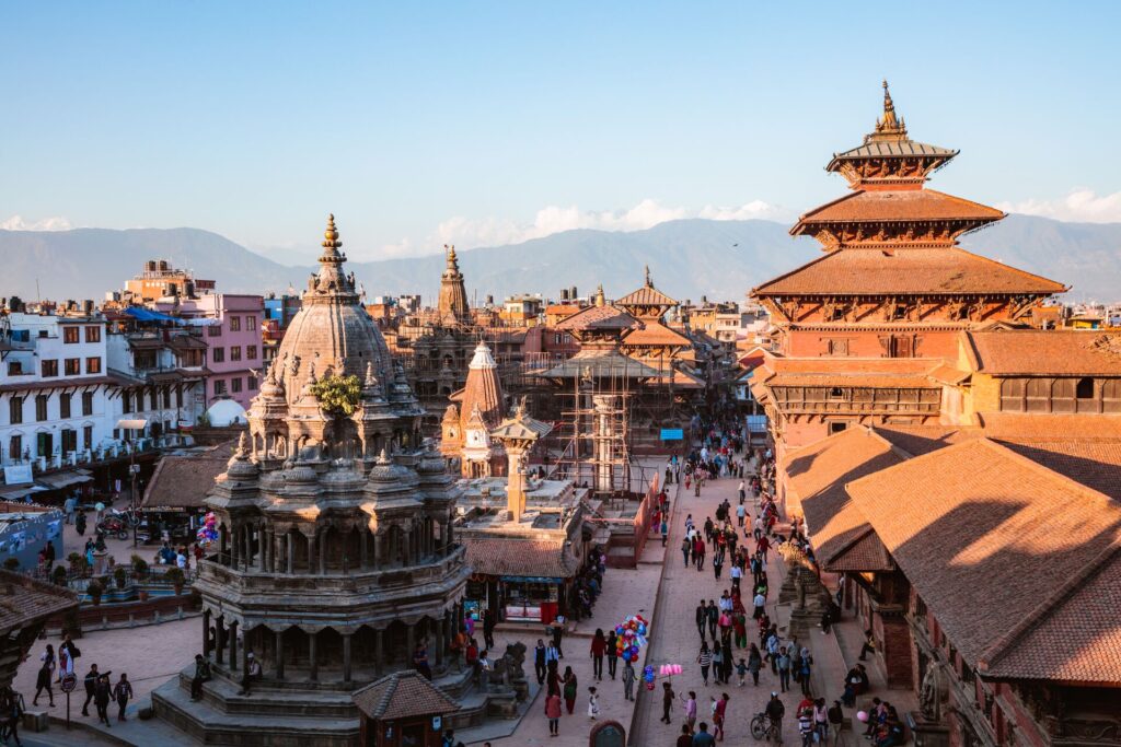 Red and brown temple roofs in Katmandu, Nepal, against a pale blue sky with mountains, and people walking around below.