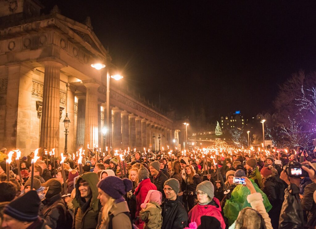 crowds with candles gather outside a large building with pillars on New Years Eve in Scotland