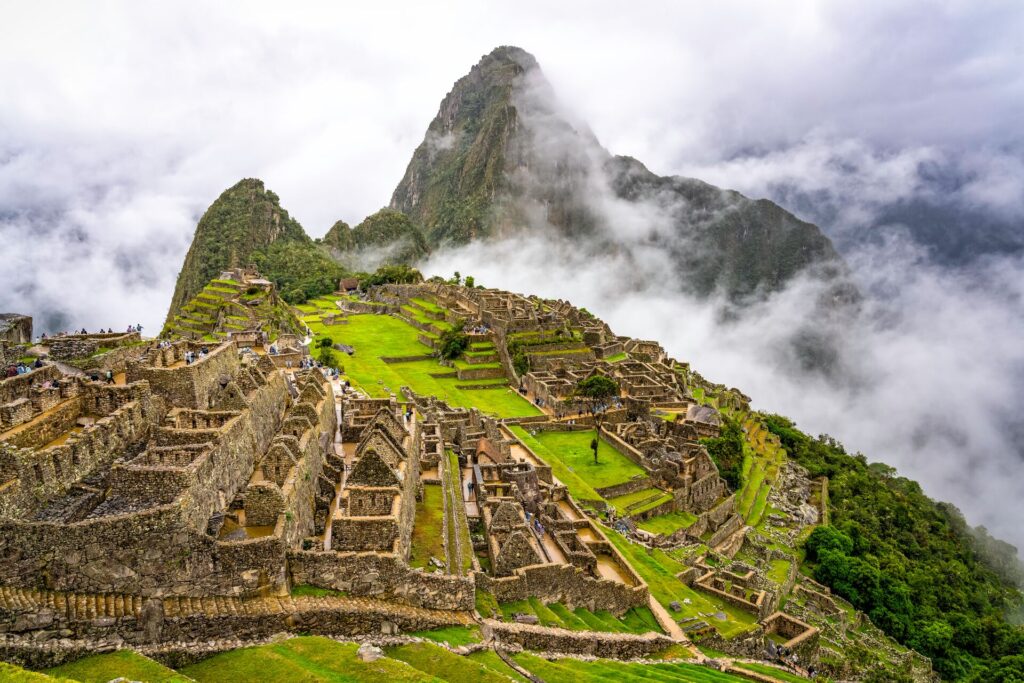 Bright green grass with brown stone terraces and buildings, on top of a green mountain in Machu Picchu, Peru, with clouds swirling around the top.