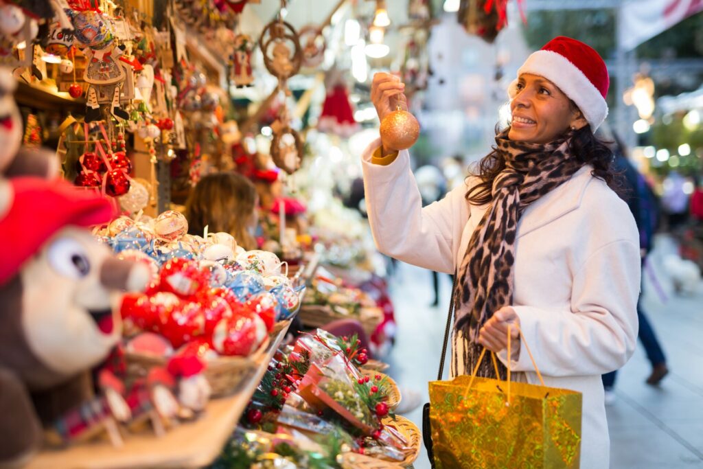 A lady looks at baubles in a festive market, wearing a red and white Santa hat and a big smile