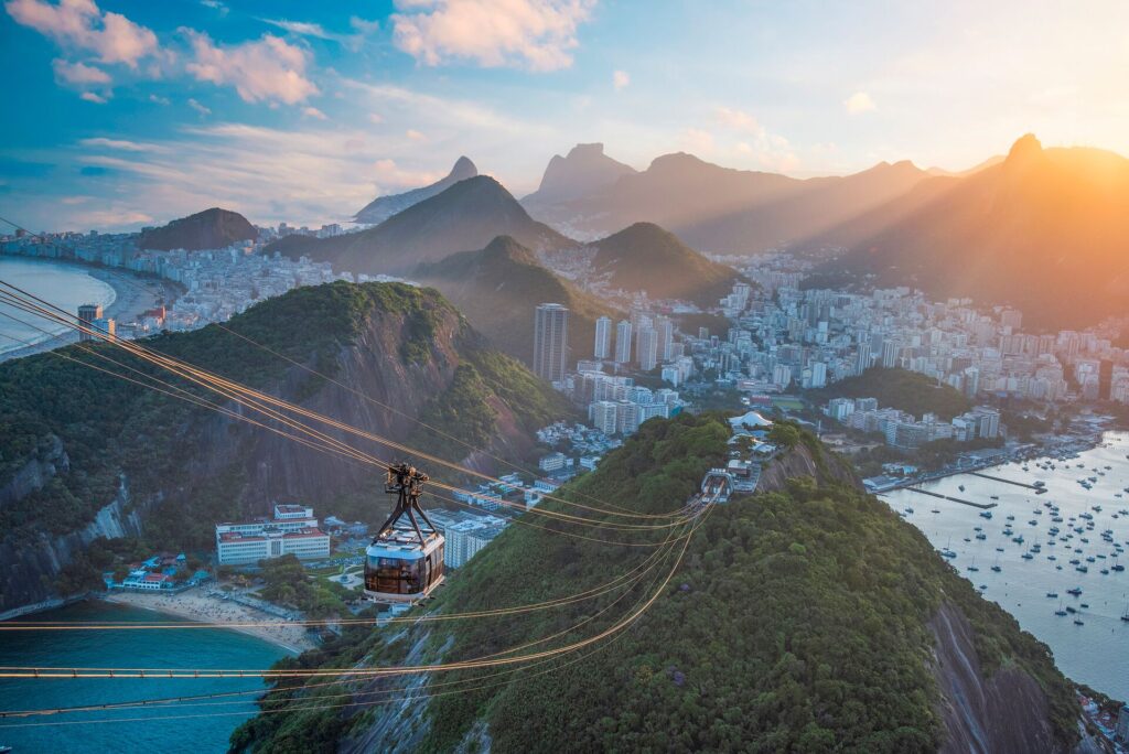 A cable car flows down to the top of a green covered mountain, with the white houses and buildings of the city .of Rio De Janiero in Brazil sitting blow, 