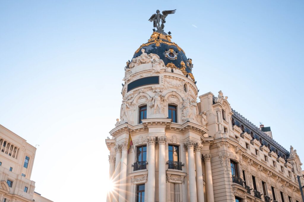 The Metropolis monument in Madrid,Spain with a blue domes roof, grey angle on the top, white facade with columns and ornate decorations, against a pale blue sky