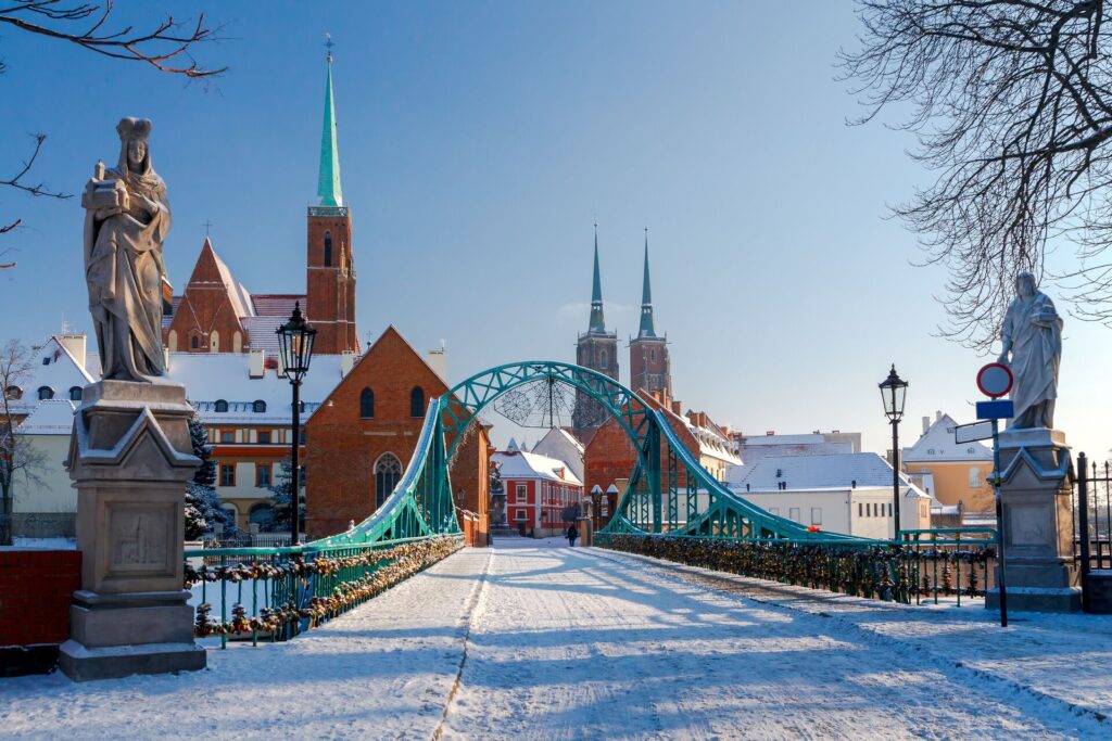 A wide bridge with green railings is dusted in snow, with statues int he foreground and church spires in the background, in Krakow, Poland