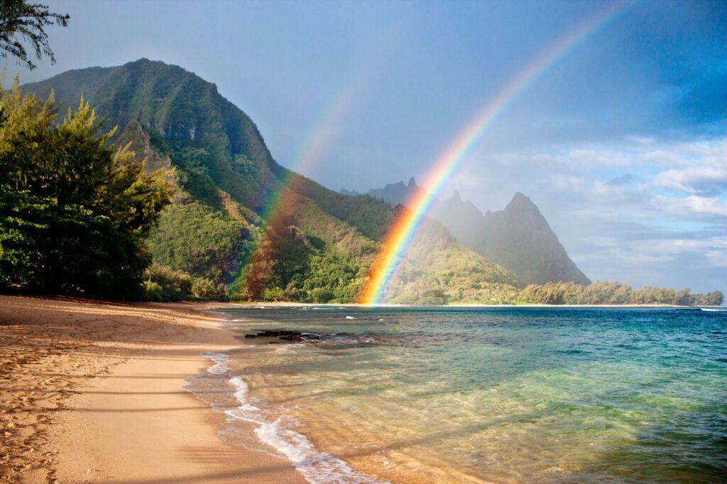 Two rainbows leap from a golden beach with blue sea and green covered mountains in the background.