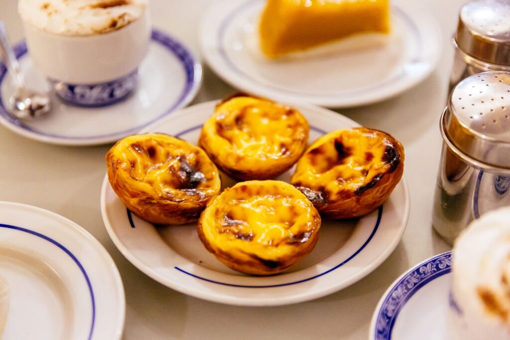 Four pastel de nata Portuguese custard tarts on a plate, with a cup of coffee and other dishes surrounding.