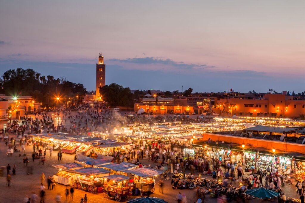 A busting Moroccan bazaar lit up at night with a tall mosque tower in the background.