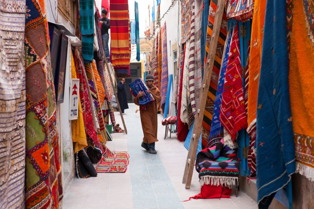 Rows of brightly coloured rugs and traditional carpets in a bazaar in Morocco, with a man walking in between.