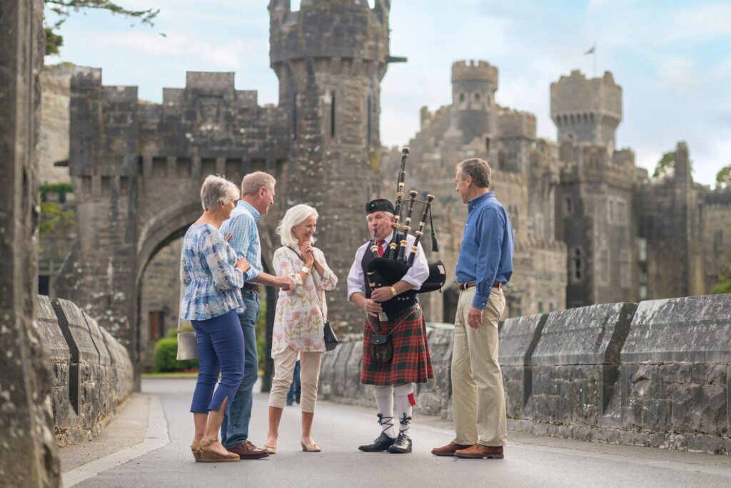 Four guests stand with a piper in traditional dress on the drawbridge of Ashford Castle, with towering grey turrets behind. 