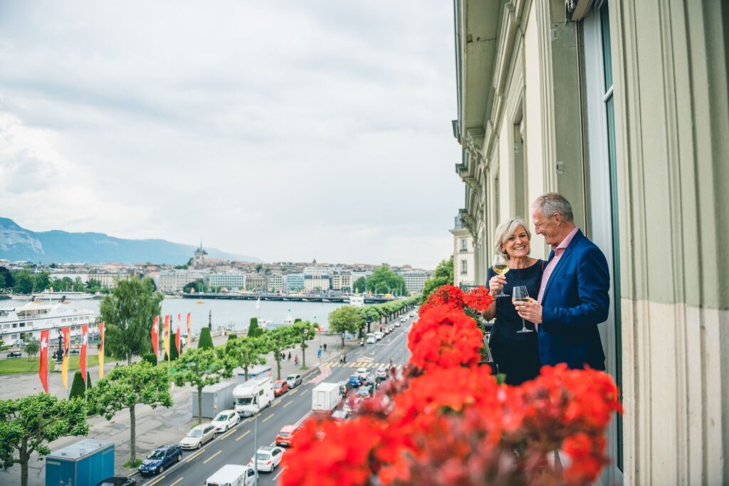 A couple share a glass of wine on the balcony of Hotel D’Angleterre, with red flowers and a view over Lake Geneva
