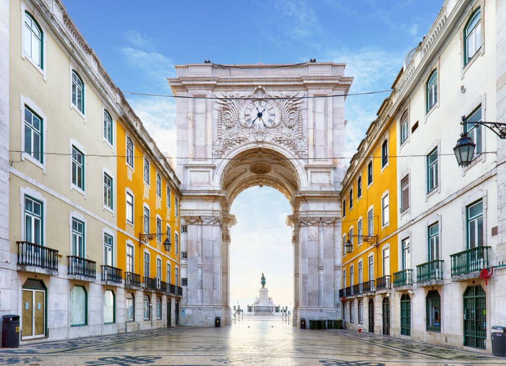 The arch at the Praca do Comercio, Lisbon, Portugal in white brick with ornate carvings, with yellow buildings either side and a tiled square in front.