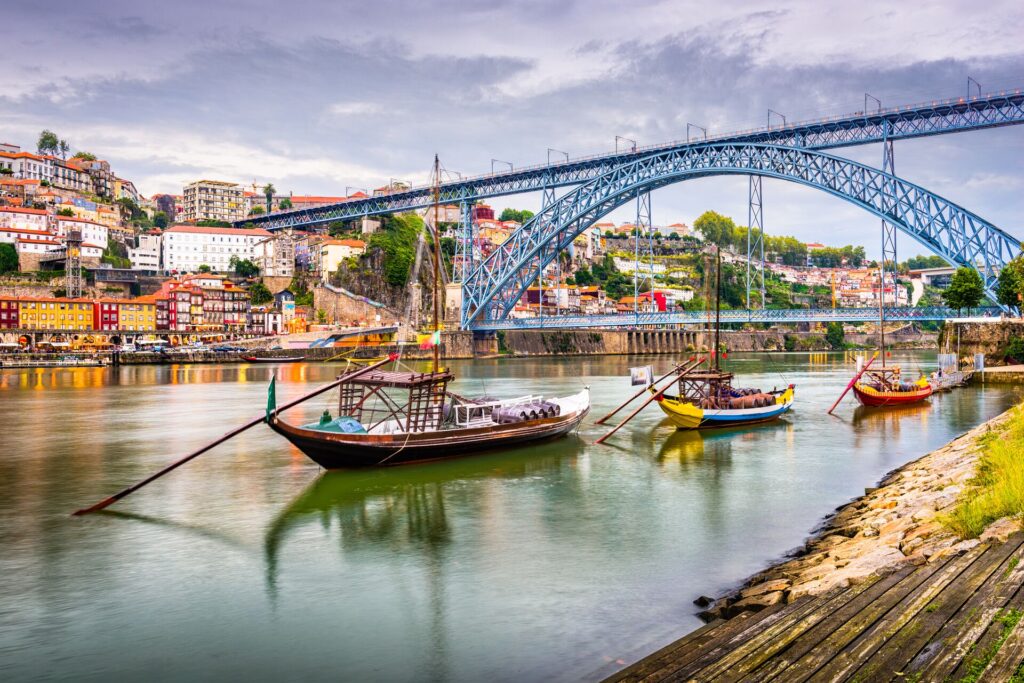 A blue metal bridge spans a wide river, with colourful houses not he banks and colourful fishing boats on the water in the foreground in Porto, Portugal.