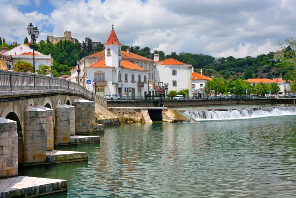 White houses with ref roofs sit on the edge of a river with an old stone bridge and a white cloudy sky.