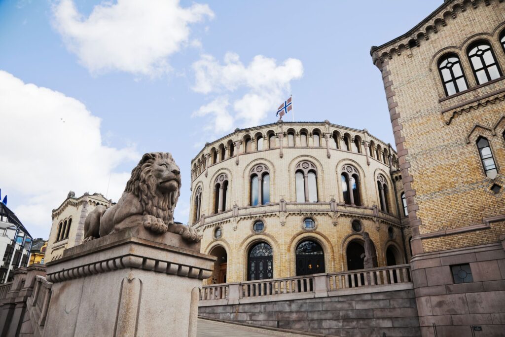 A stone lion guards the Norwegian Parliament building on Oslo, a round structure with high arches and a Norway flag flying from the roof.