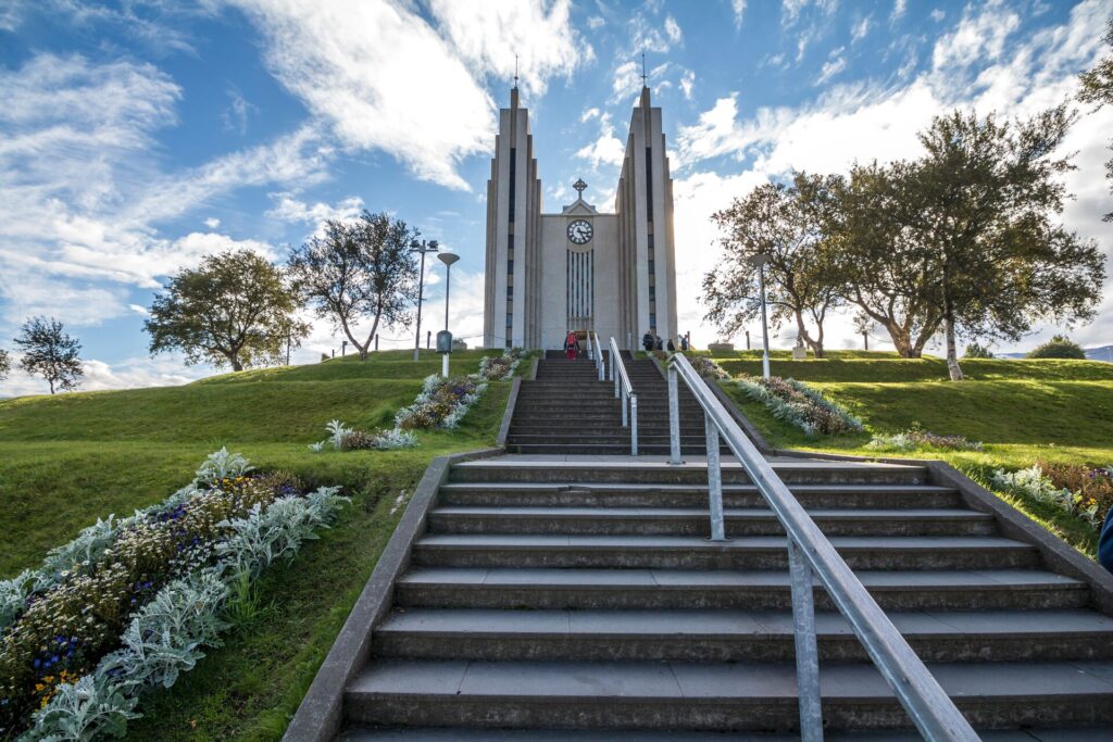 Wide grey steps lead up to a modern church with two turret either side and green laws with flowers in Akureyri, Iceland.