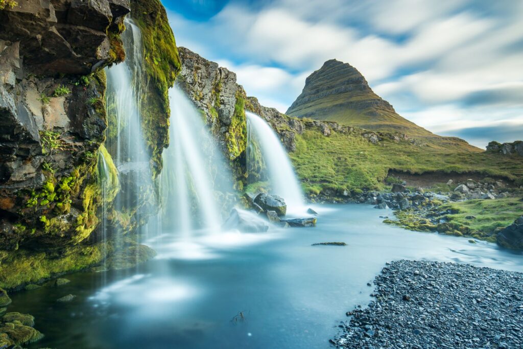 Wanterfalls cascade into a blue lake with green mountains behind, in Iceland.