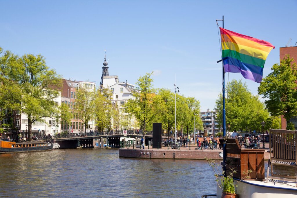 A Rainbow Pride flag flies on a flagpole alongside a canal, with green trees and white buildings in the background, in Amsterdam.