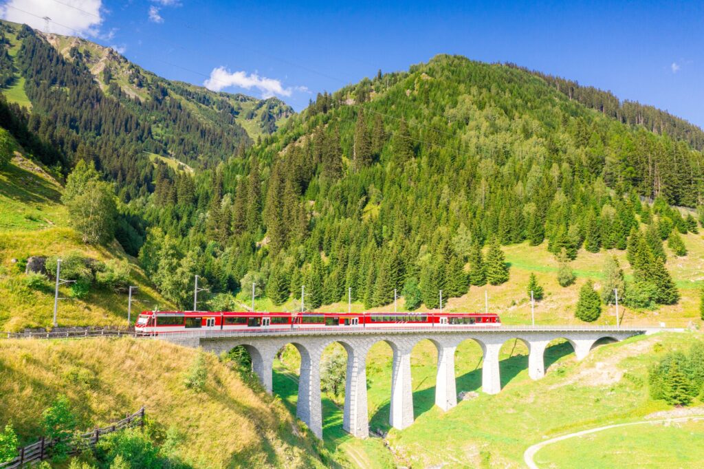 Switzerland’s bright red Glacier Express train travels over a white stone aqueduct with bright green hills behind and blue sky.