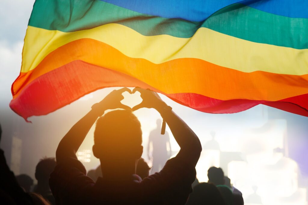 A silhouette of a person making a heart sign with their two hands, with a rainbow Pride flag flying above.