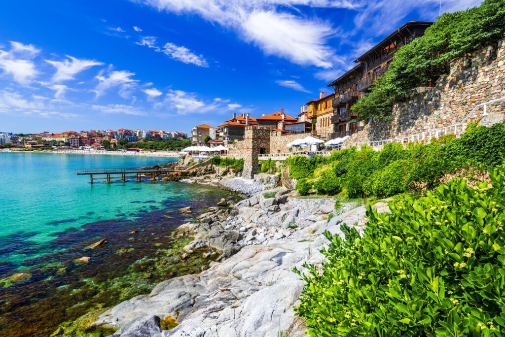 A town of red roofed brick houses sits alongside a beach and green sea, with lots of bright green foliage.