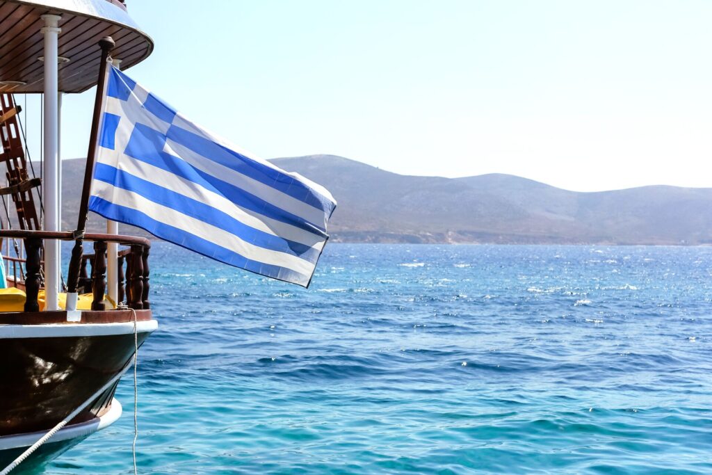 The blue and white Greek flag flies from the bow of a ship sailing on blue sea water.