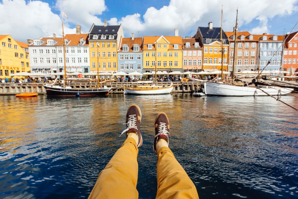 Colorful houses in blue, yellow, pink and white with boats and a dark blue river front, and the photographers legs in view.