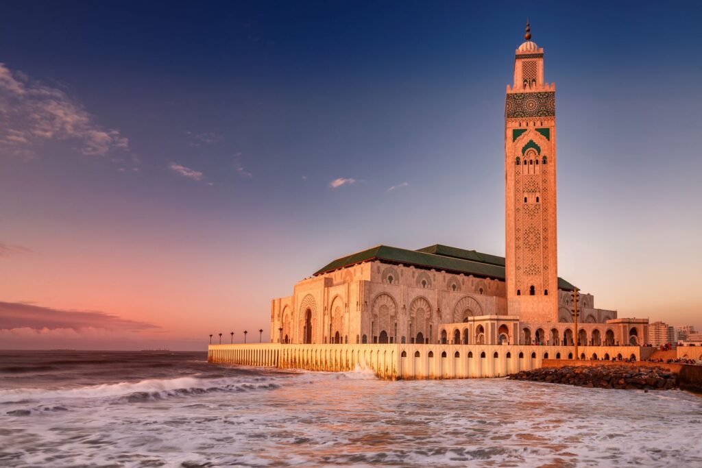 The Hassan II Mosque in Casablanca with its tall turret and white buildings, pink in the sunset.