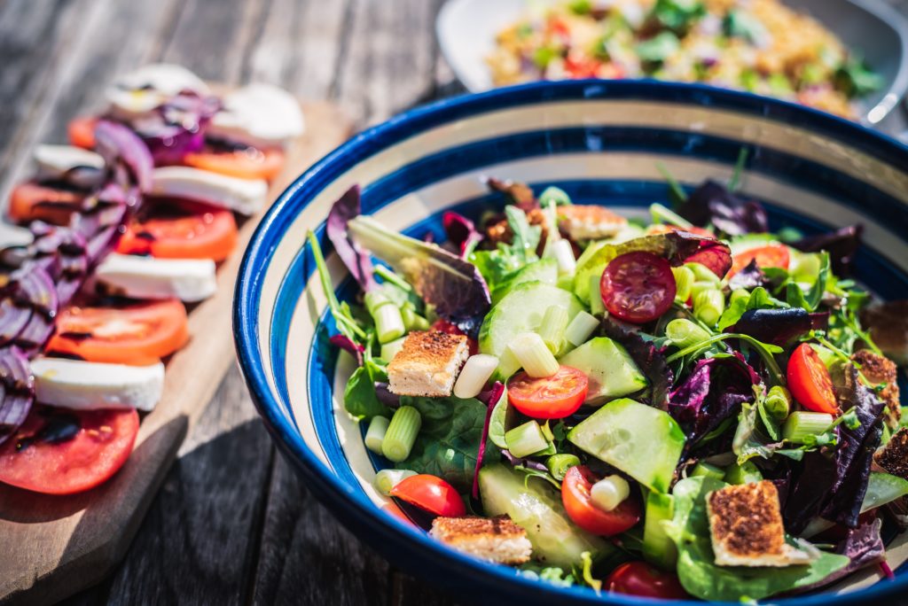 A blue and shite striped sand bowl filled with green cucumber, red tomatoes and other colorful vegetables and salad leaves.