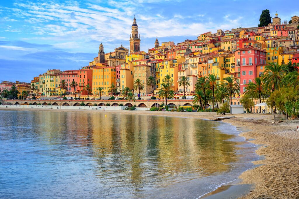 Bright yellow, range and pink houses on the French Riviera with blue water in front.