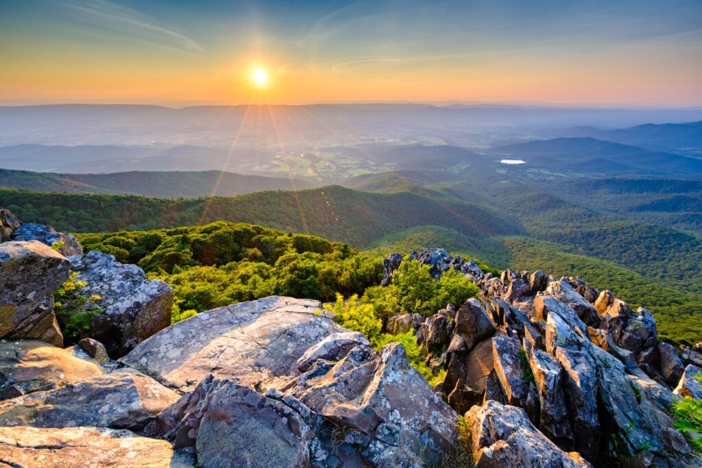 The sun shines over a vast green forrest landscape, with jagged rocks in the foreground in Shenandoah National Park in the USA.