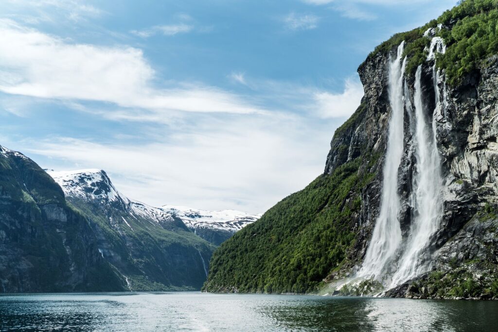 A deep blue water fjord with towering mountains either side and cascading waterfall in Norway.