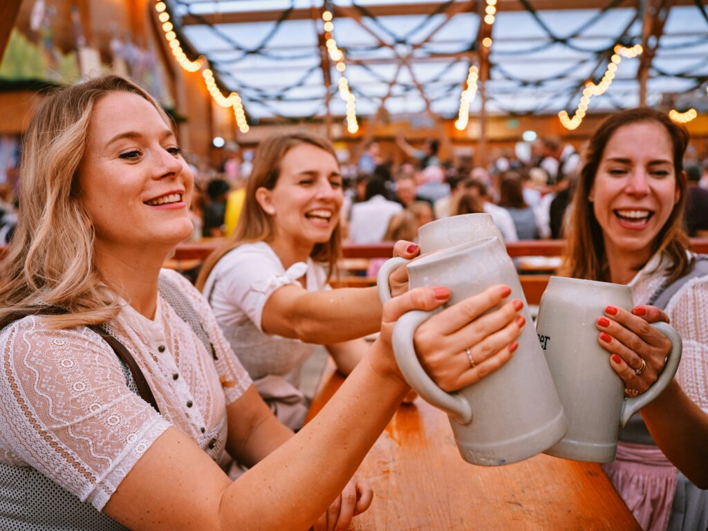 Three young women clink large beer casks and smile, with crowds in the background at Oktoberfest.