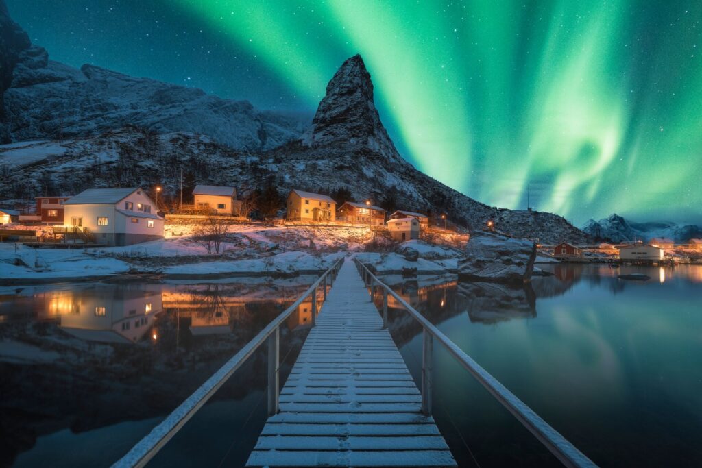 The Northern Lights dance bright green above a snow covered village lit up at night in Norway.