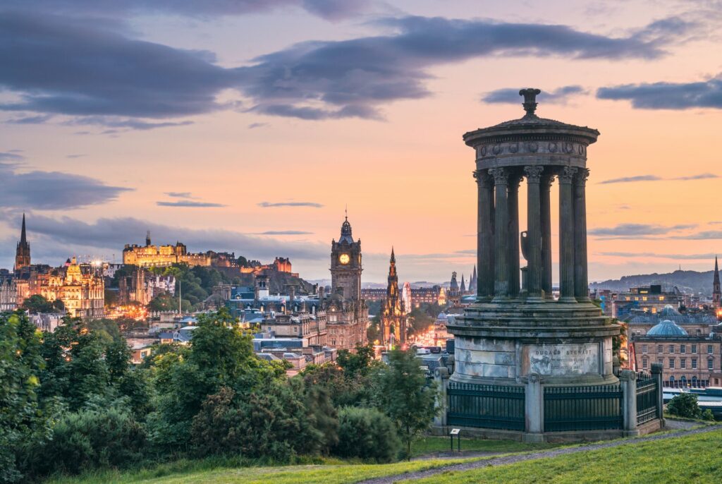 Cityscape pf Edinburgh, Scotland, with a stone monument, green parkland and the city behind with a large clock tower.