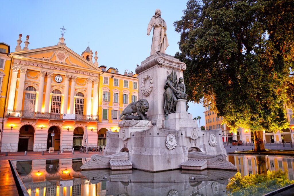 A white stone statue with lions and people, with a large, yellow palace faced behind, lit up in the evening fall sun.