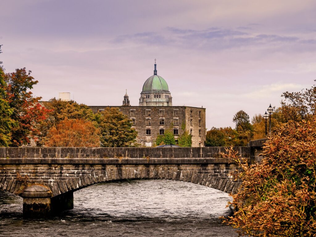 A stone bridge spans a river in Galway, Ireland, with crimson fall foliage and a large, green domes building in the background.