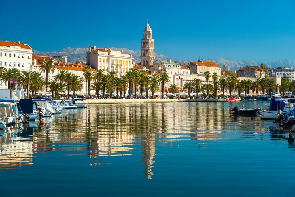 Large white buildings with orange roofs flank the water's edge in Split, Croatia, reflecting the water with bloats on either side.