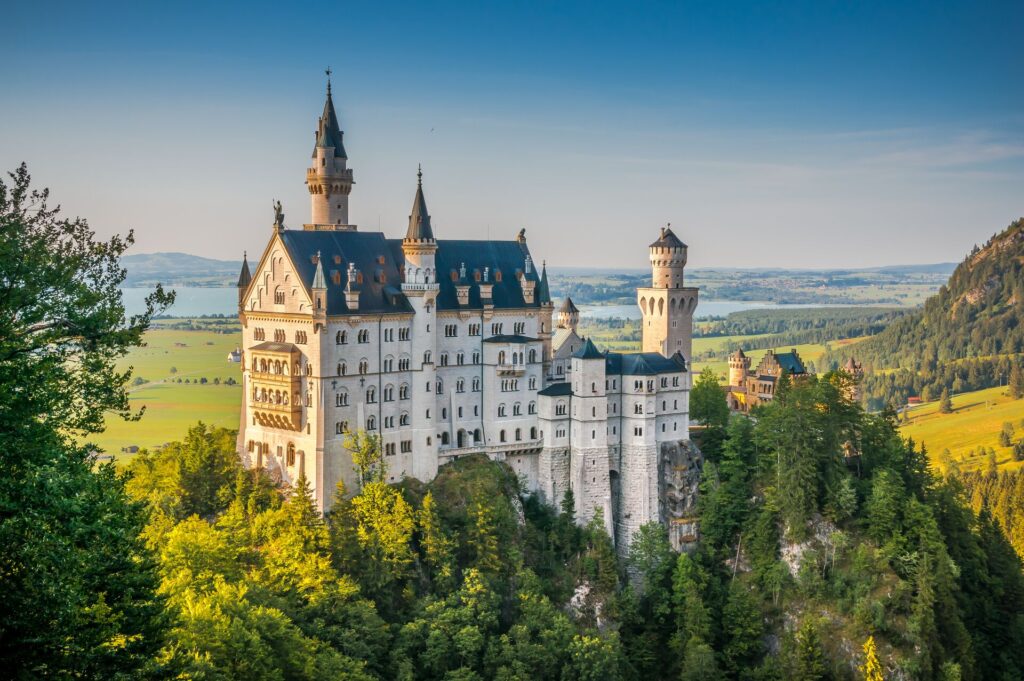 Fairy-tale Neuschwanstein Castle, white with turrets overlooking green lands and a lake.