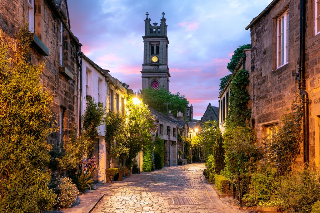 An alleyway in a scottish city, the sky is violet and the cobbled street curves away into somewhere unknown. Charming cottages line the alley, each with lush foliage lining their facades. A clock tower stands erect in the background.