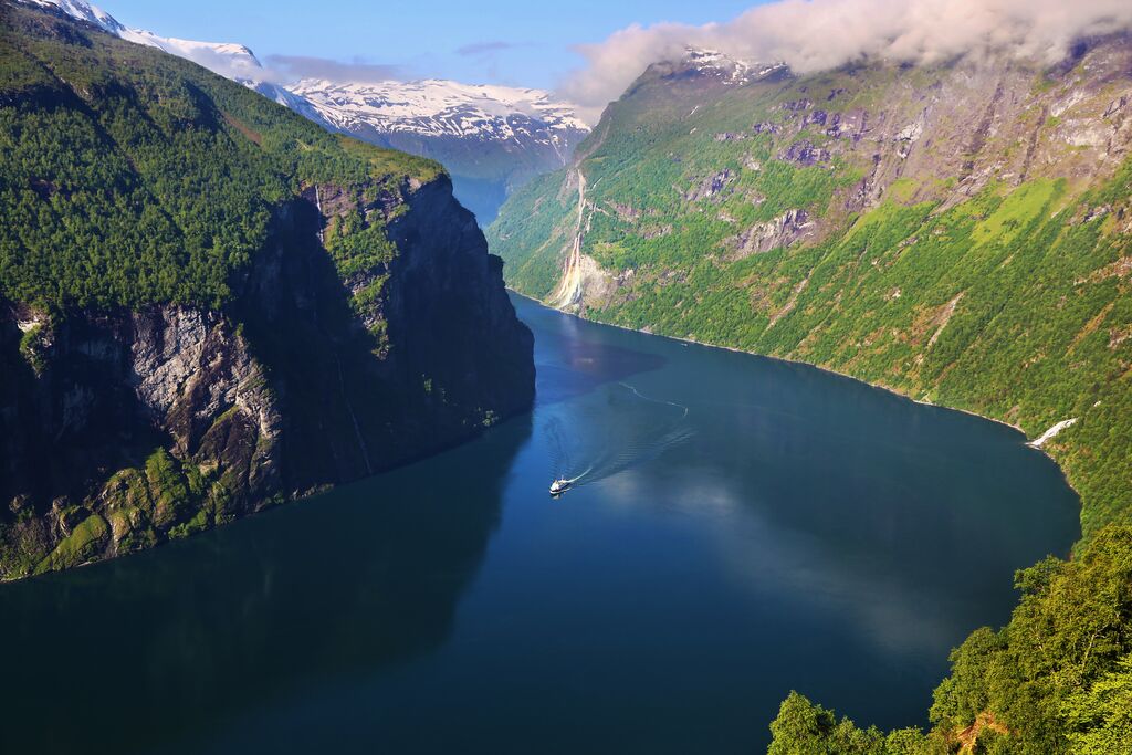 A huge fjord curves around a valley, with a boat cruising through its waters and snow-capped mountains making up the background. 
