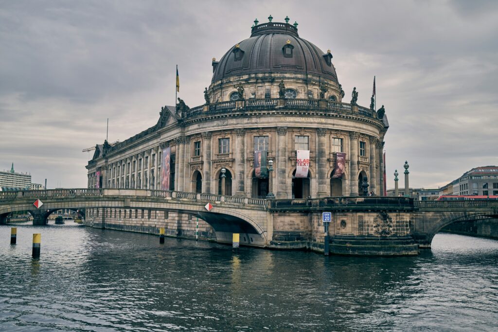 A grey museum building is shown on the water with bridges leading to it in Berlin.