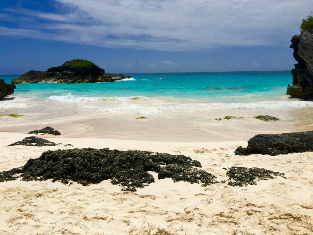 Azure water laps onto a faintly pink beach with black rocks on the beach.