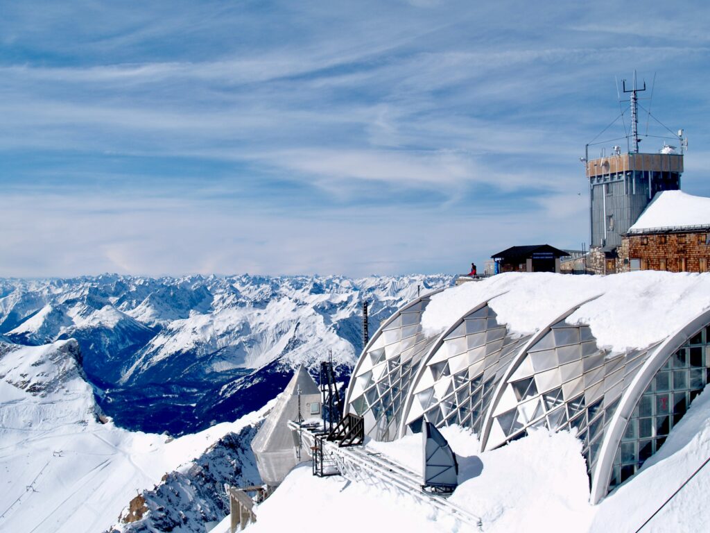 A snowy mountains summit with a viewing building, with a view of=ver mountains and bright blue sky.