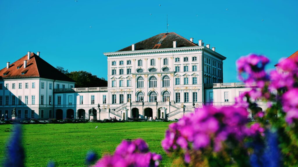 Impressive white facade of Nymphenburg Palace in Germany with green lawns and purple flowers in front