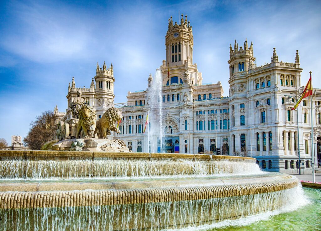 The white and gold Cibeles Fountain In Downtown Madrid, Spain is shown with a n ornate fountain in front