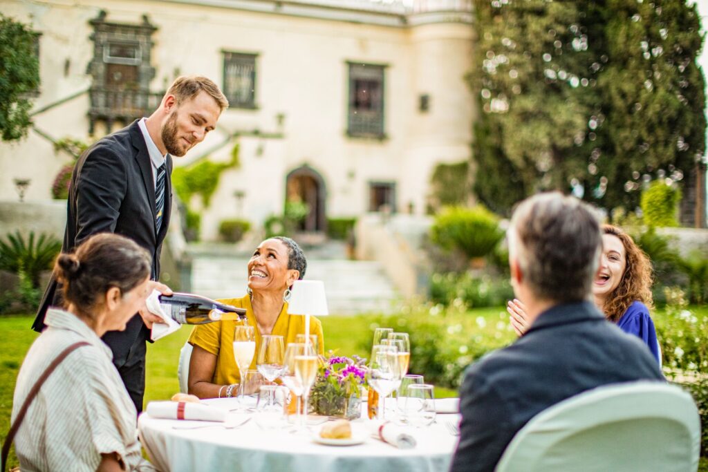 A waiter pours wine for four adults sitting around an outdoor dining table
