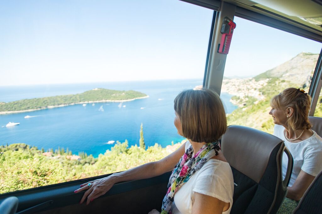 two mature women look out the window of a coach towards the blue waters of a mediterranean coastline