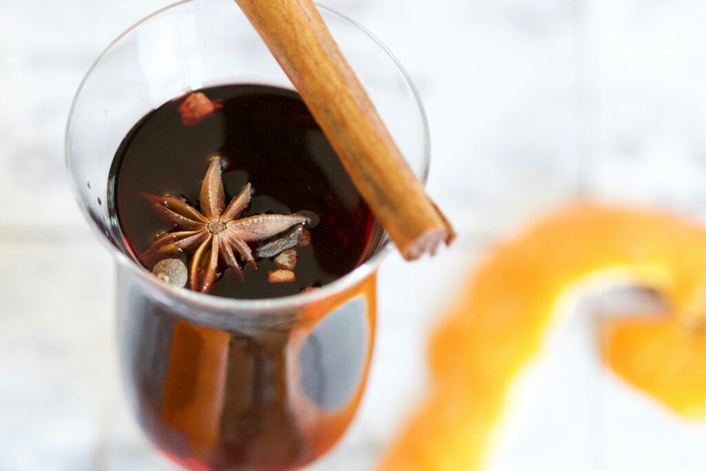 A glass of dark red mulled with with spices floating on the surface, next to a bright strip of orange rind