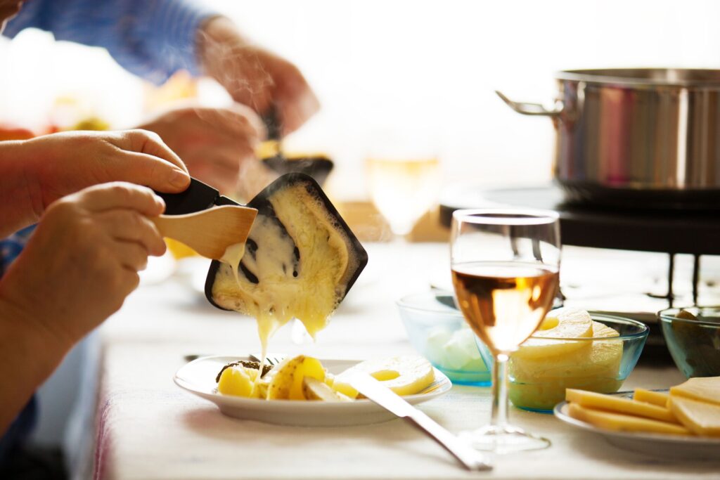 Hands are shown scraping raclette cheese on to a plate, from a small tray on which it is melted. 